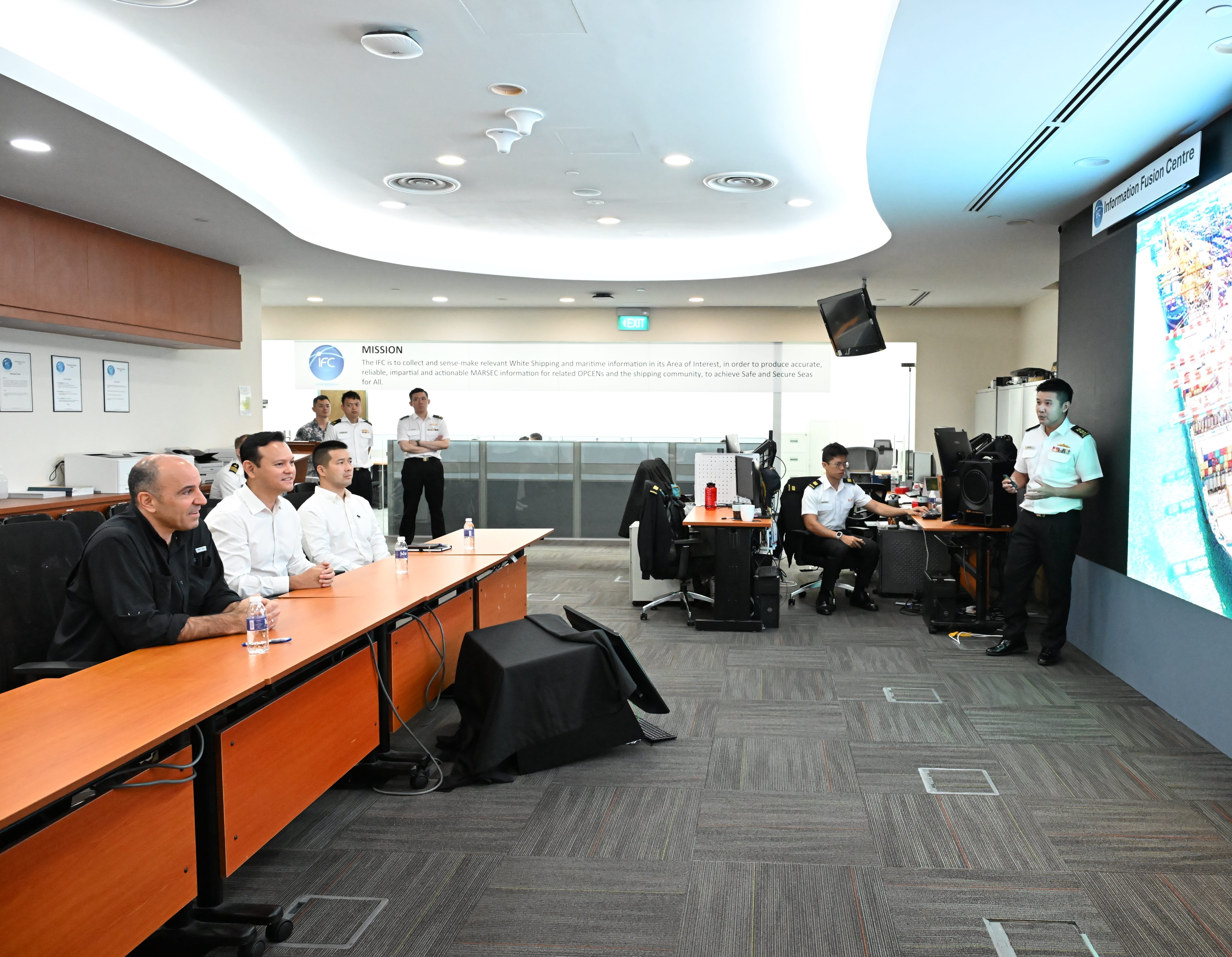 People in office setting. Presenter in uniform stands by screen showing ship. Meeting table with attendees.